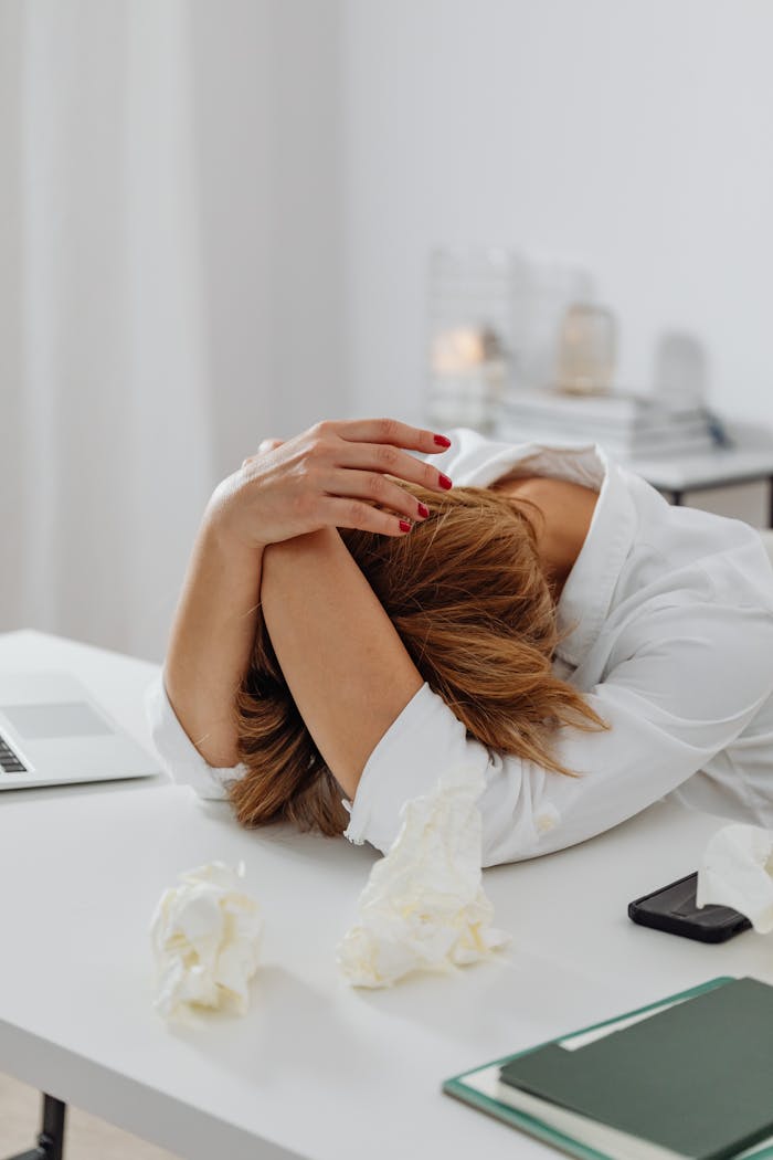 A woman in distress sits at a desk with head in hands, surrounded by tissue papers.